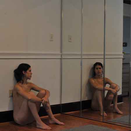 Photograph of a figure sitting on the ground, legs bent and arms draped over their knees. The figure is holding their right wrist with their left hand as they stare into a four-part mirror, their reflection staring back at them.