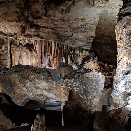 Photograph of a well-lit cavern. The foreground is mostly rock walls and some stalactites are in the far background.