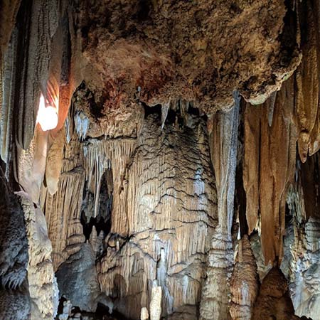 Photograph of a cavern ceiling. The center foreground is a large uneven rock formation with a rough texture surrounded by drapery formations: one of these formations is lit from within to show towel-like striations within the formation. The background has a variety of flowstone formations that look like frozen waterfalls.