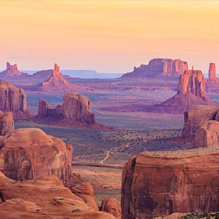 Photograph of a desert landscape accented with red-orange buttes and mesas. The image is viewed from above, with portions of rock formations visible before giving way to the valley below. The natural light casts a purple hue across the sparse landscape.