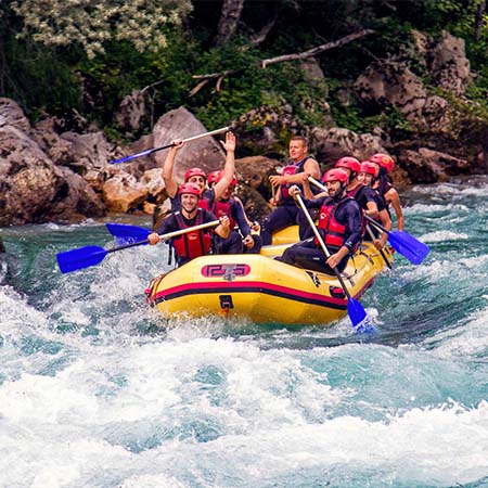 Photograph of a group of whitewater rafters. The water is tumultuous, mostly white and light blue with a few spots of darker water. The rafters are aboard a yellow inflatable raft, wearing red helmets and life vests, and using blue oars. Behind them, some greenery is visible along the rocky shoreline.