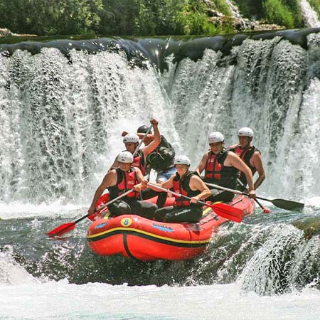 Photograph of a group of whitewater rafters about to go down a short waterfall drop in a dark green river. The water below is white with foam, and behind them is a much larger waterfall, about twice the height of the rafting group, flowing from crystal-smooth water to tumultuous white foam. The rafting team is in a red boat with matching oars and life jackets, wearing white helmets.