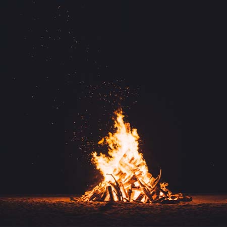 Photograph of a campfire at night. Sticks have been positioned in a cone-like structure, and the fire blazes in a similar, conical, shape, blown ever so slightly to the left - orange sparks being thrown off and picked up by the wind. The center of the fire burns white and the edges of the flames are a light orange-yellow.