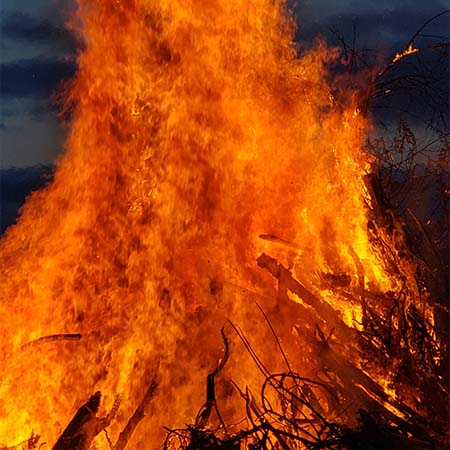 A close-up photograph of a massive bonfire. The kindling is only visible as a black silhouette against the red-orange flames that billow above the top of the image frame.