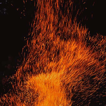 Photograph of a dense, upward, explosion of orange-yellow sparks. The sparks appear as lines of light in the darkness, buffeted by the breeze before traveling upward.