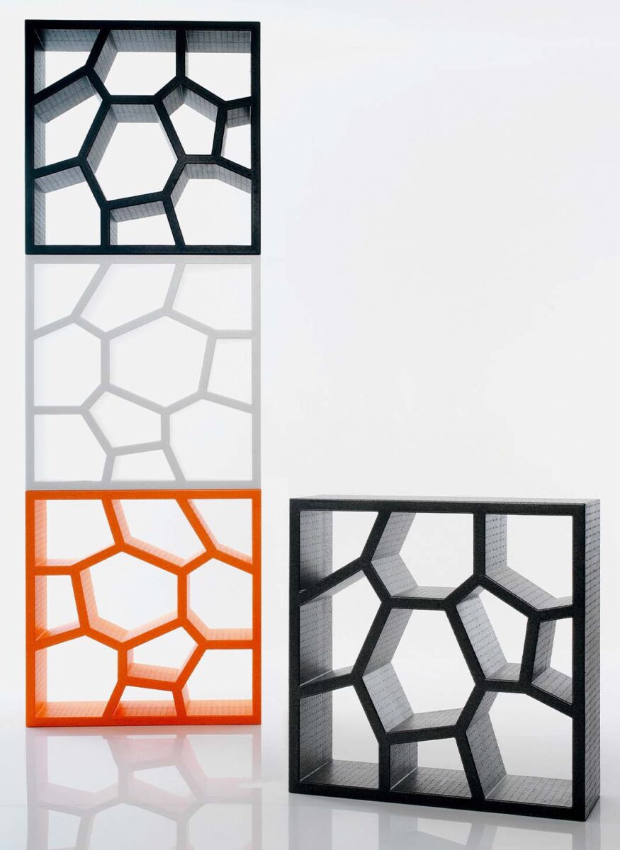 Square, plastic, stacking storage units with irregular geometric shapes creating the storage surfaces within. The left of the image shows three of the units (in black, white, and orange) stacked together, and the lines creating the irregularly shaped storage nooks flow seamlessly from one unit to the next. The right of the image shows a single black unit at a slight angle, demonstrating the depth of the shelves. 