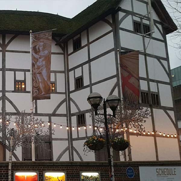 Photograph of a portion of the Globe Theatre’s exterior: its high walls are defined by vertical and horizontal timbers, with occasional diagonal supports, creating a grid-like pattern that is filled in with whitewashed plaster.