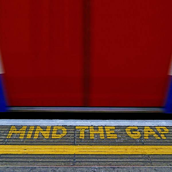 Photograph of the “Mind the Gap” slogan painted on the edge of a London tube station platform. The yellow text on the dark gray concrete is in perfect focus, while the closed bright-red doors of the train car are blurred in motion.