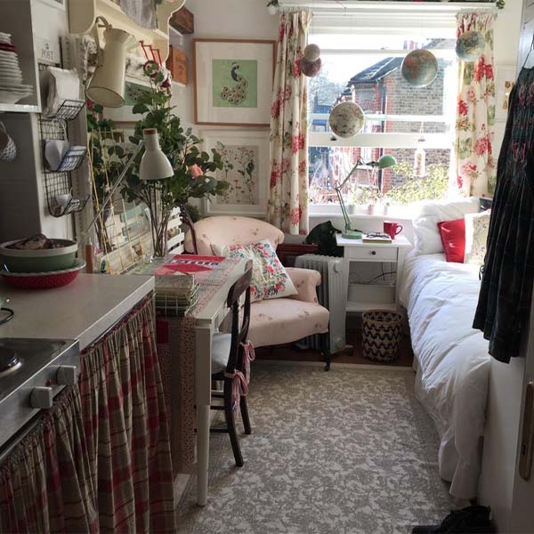 Photograph of a small London apartment The kitchen is barely visible in the left of the image and butts right up against a small table with a single chair. In the corner behind the table is an armchair; to its right is an electric heater that is also to the left of a small bedside table that is pressed right up against the bed that takes up the remaining space.