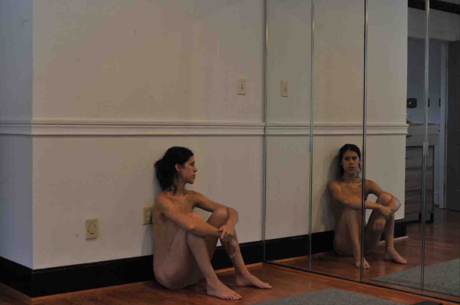 Starting image: Photograph of a figure sitting on the ground in a dimly lit room, their legs bent and arms draped over their knees. The figure is holding their right wrist with their left hand as they stare into a four-part mirror, their reflection staring back at them. The figure leans against a plaster wall with molding a few inches above their head. The molding in the reflection is seen to travel further to a turn in the wall, beyond which is a dresser and an open door.