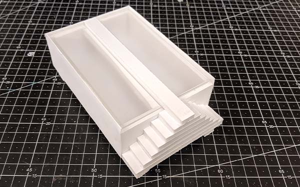 Side 1 of foam core table model D, predominantly rectangular, the short edge of the closest size has a small, two-sided, staircase leading up to a “glass” tabletop with a line of foam core running down the center.