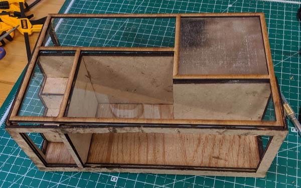 Side 1 of a laser-cut wooden table model. It’s fully framed with irregularly shaped rooms formed by four “walls” that run perpendicular to each side. The rightmost “room” in this view has a small staircase and the right room is a fat “L” shape. The glass top sits below a frame that matches the layout of the “walls” below, and the top right square section of the glass is fogged.