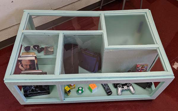 Side 1 of a full-scale foam table model. The layout is identical to that of the laser-cut wooden table, fully framed rectangle with irregular walls running perpendicular to the edge of the table,  and has objects placed within it. The “stairs” have sunglasses, keys, and books stored on it and the “L” shaped section has games and knicknacks.