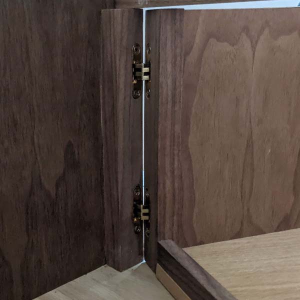 A closeup image of the pull-out cabinet and its hinges. The cabinet walls are made of walnut and the base of the same whiteoak as the table base. The white oak base of the pullout is framed with solid walnut to prevent items from falling when opened, and the same solid walnut is mounted to the walnut wall of the “room” and the pullout to embed the no-show hinges.